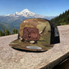 Camouflage cap with a deer patch on a stone ledge with mountains in the background