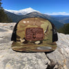 Camo hat with a deer logo on a rocky surface with mountains in the background
