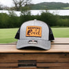 fishing hat with 'Keeping It Reel' patch on a wooden surface with a scenic background.